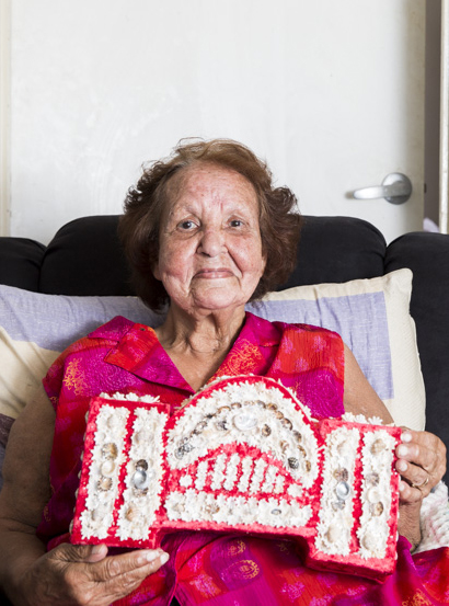 Smiling portrait of an old woman holding a model of the Sydney Harbour Bridge covered in shells.