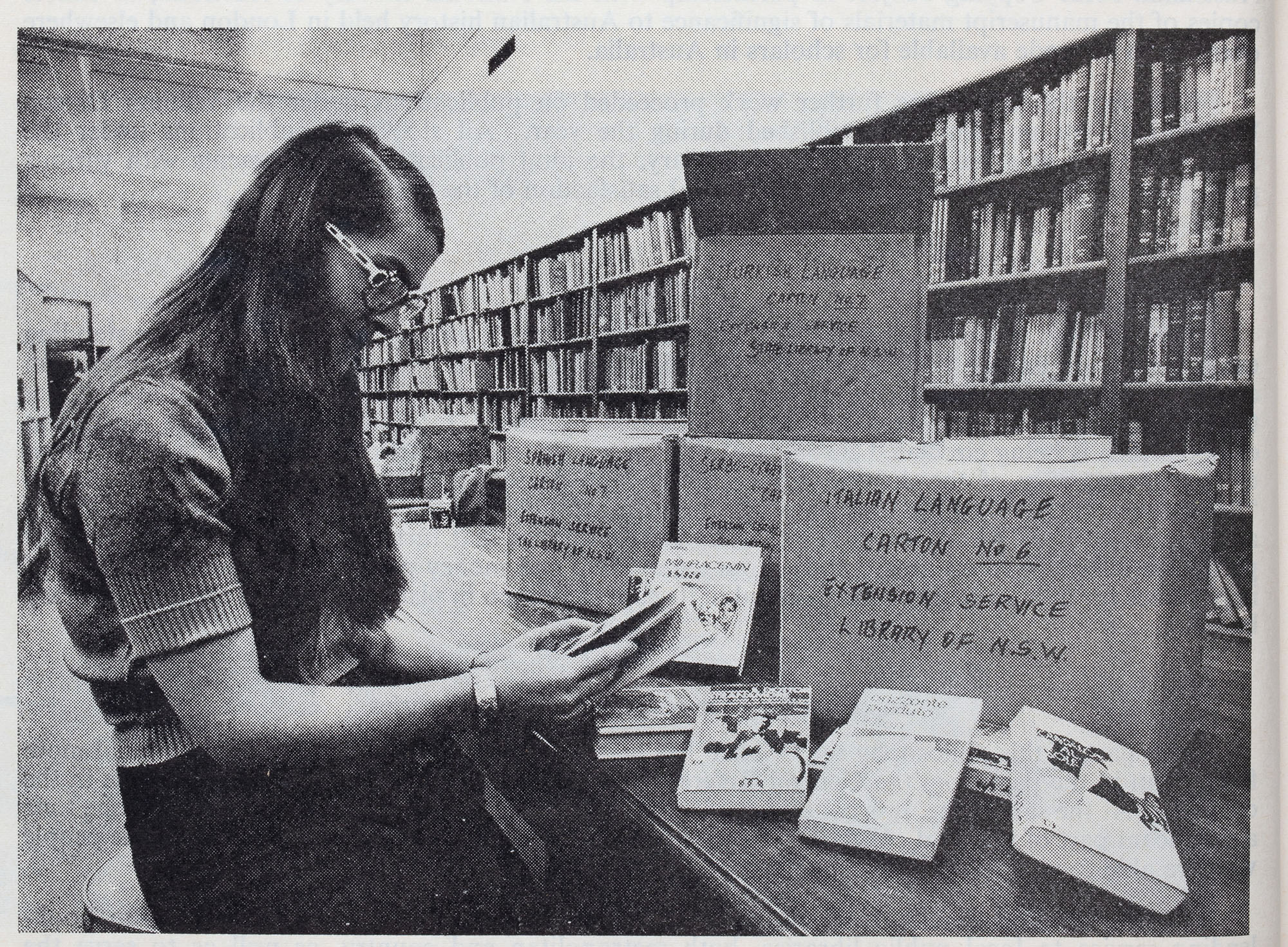 Black and white photo of a woman at a desk with boxes and books