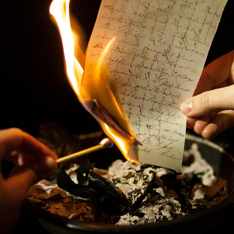 Hands holding a lit match to a burning piece of paper.
