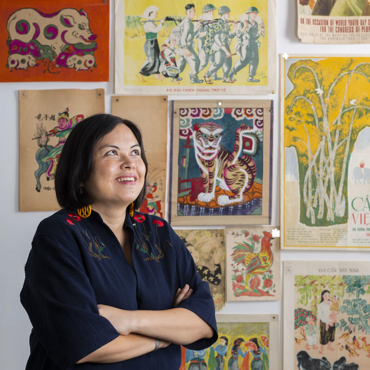 A woman stands in front of a wall of Vietnamese art posters, smiling.