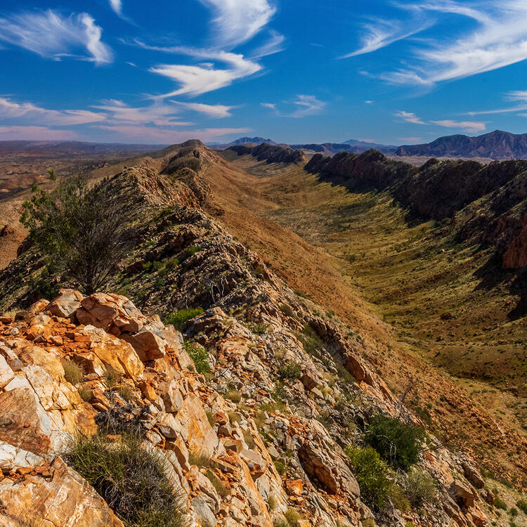 Ridge of a rocky mountain peak stretching out into the distance with blue sky above. Wisps of clouds dot the sky.