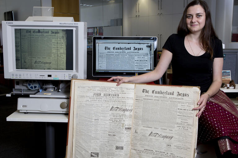 Girl holding up newspaper with image on computer next to her