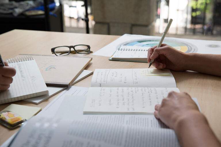 Notebooks and paper on table with arms writing on them.