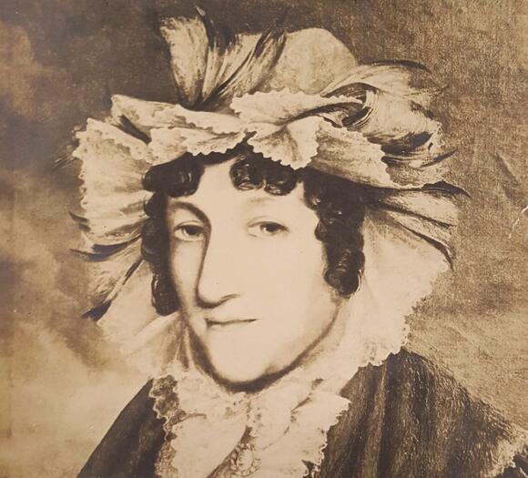 A portrait of a woman wearing a very ruffled bonnet, ruffled blouse and coat.