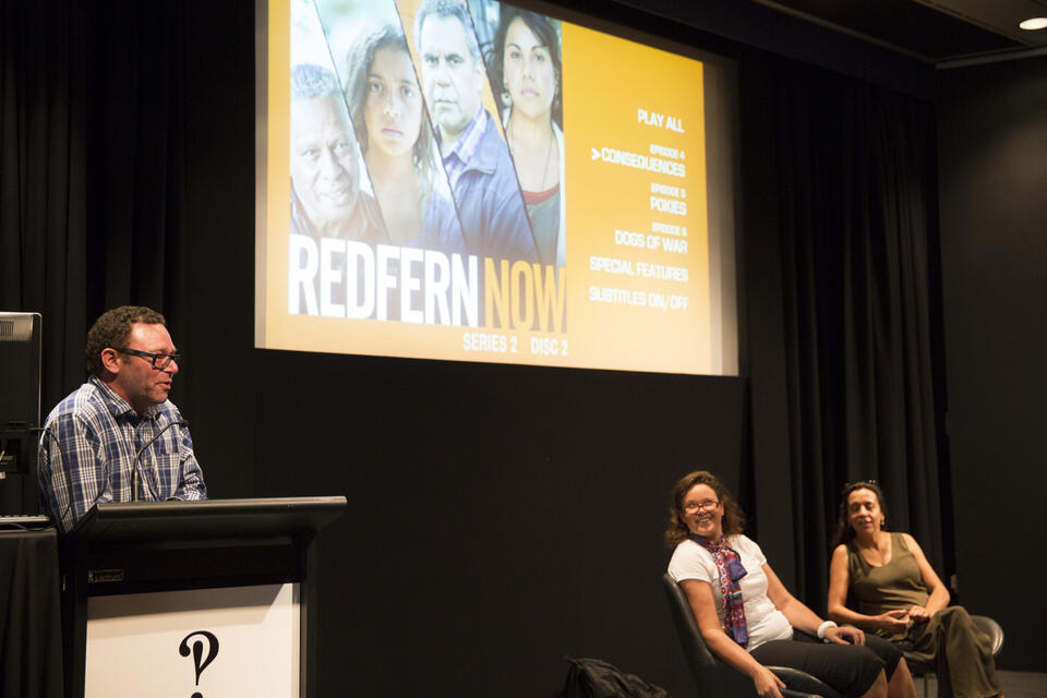 2 women sit down on a stage and with a screen behind. Another man is speaking at a microphone 