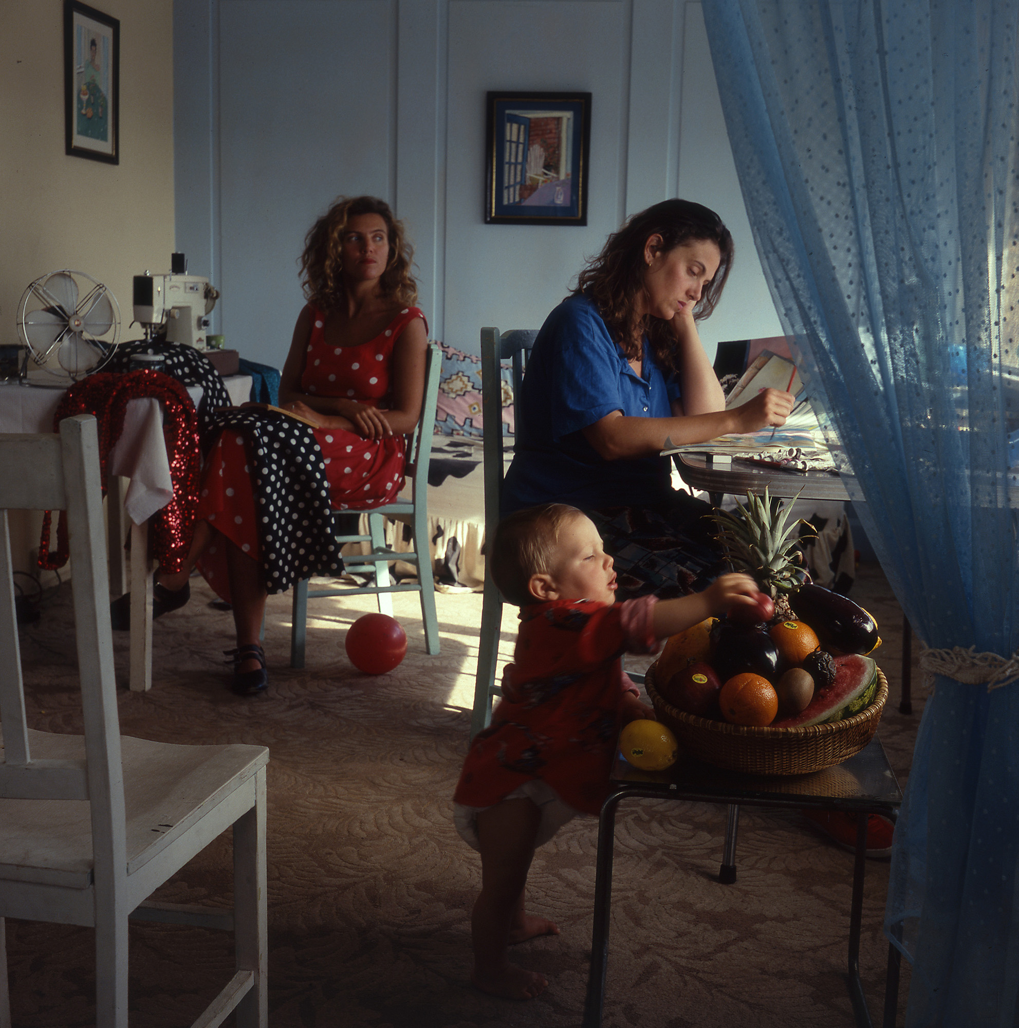 Photograph of two women sitting in a room and a baby stands in the foreground.